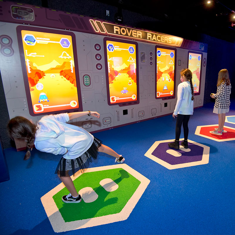 At Rover Racers in Planet Play at Kennedy Space Center Visitor Complex, explorers use their bodies to control Mars rovers, moving left and right to collect samples or jumping to avoid obstacles.