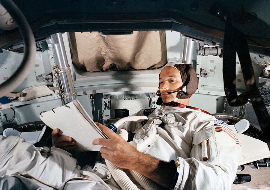Michael Collins laying down, observing a checklist in a simulator of the command module.