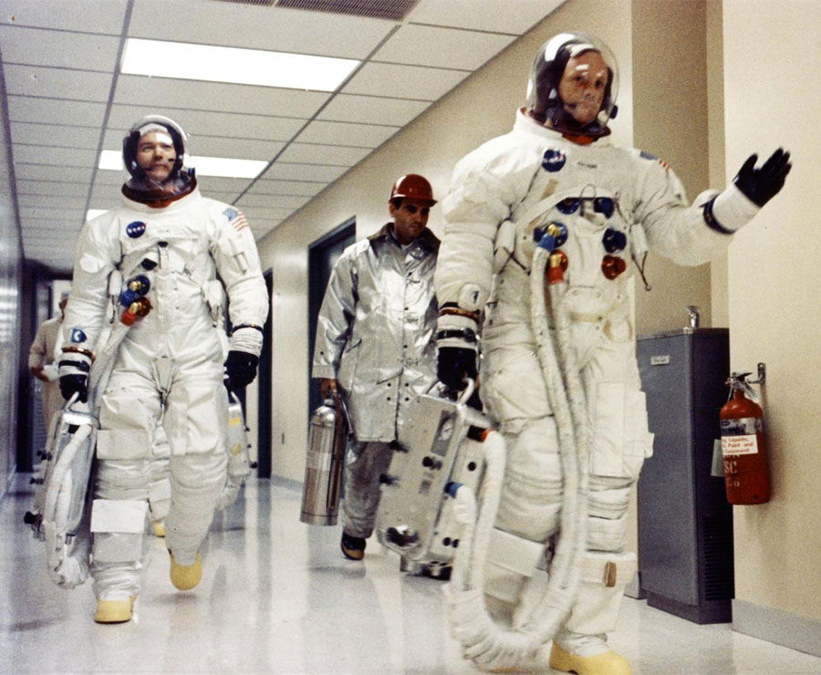 O comandante da Apollo 11, Neil A. Armstrong, acena para os simpatizantes no corredor do Manned Spacecraft Operations Building enquanto ele, Michael Collins e Edwin E. Aldrin Jr. se preparam para serem transportados para o Launch Complex 39A para a primeira missão tripulada de pouso lunar