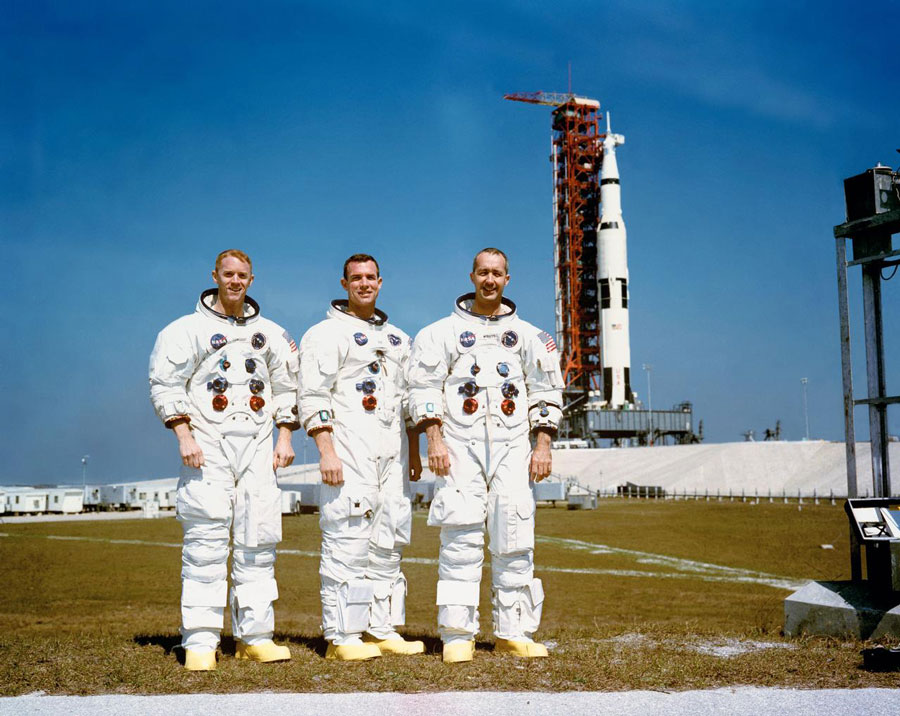 Estes três astronautas são a tripulação principal da missão espacial Apollo 9, orbital na Terra. Da esquerda para a direita, estão Russell L. Schweickart, piloto do módulo lunar; David R. Scott, piloto do módulo de comando; e James A. McDivitt, comandante. No fundo à direita está o veículo espacial Apollo 9 na Plataforma A, Complexo de Lançamento 39, Centro Espacial Kennedy (KSC).